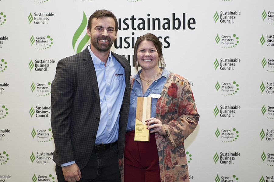 Man and woman smiling in front of branded backdrop while holding paper award.