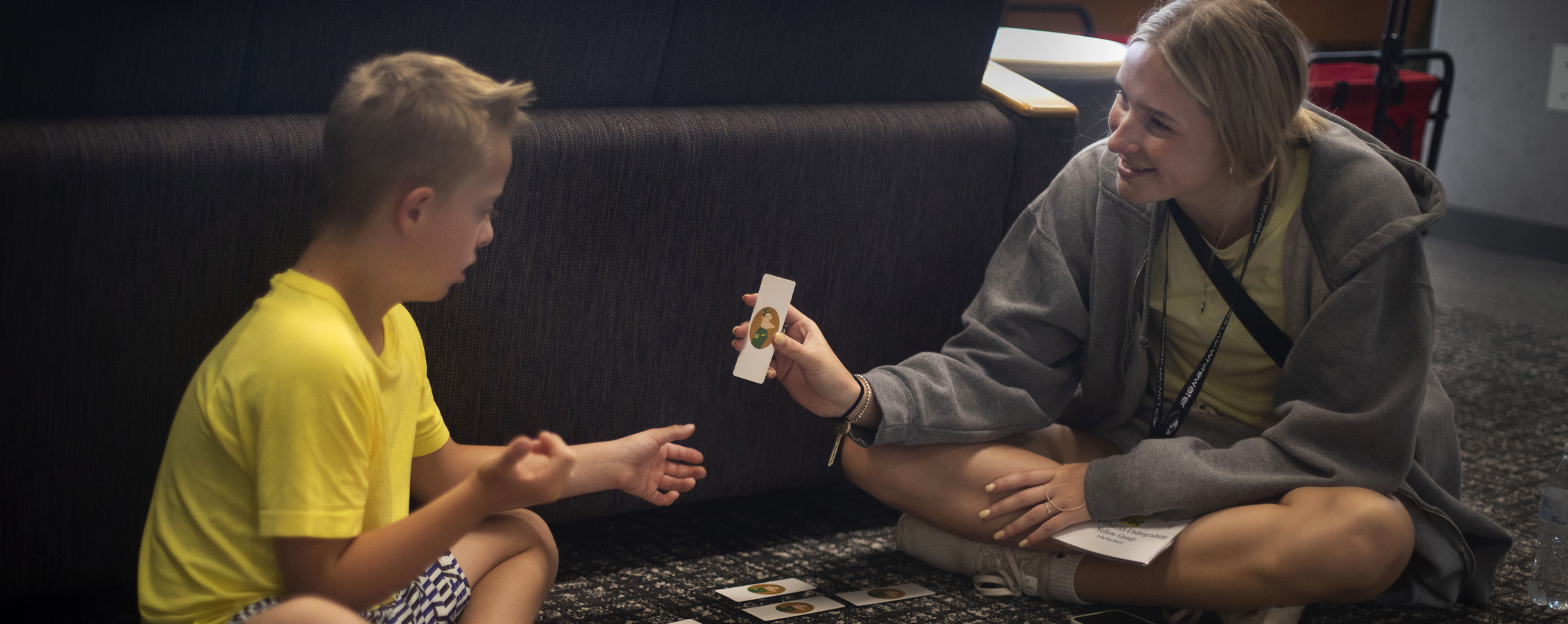 A student sits on the floor with a child as they work through flashcards.