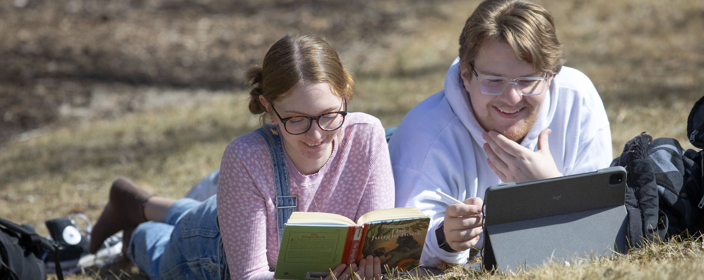 Two students lay on their stomachs in the grass and read books and work on an iPad.