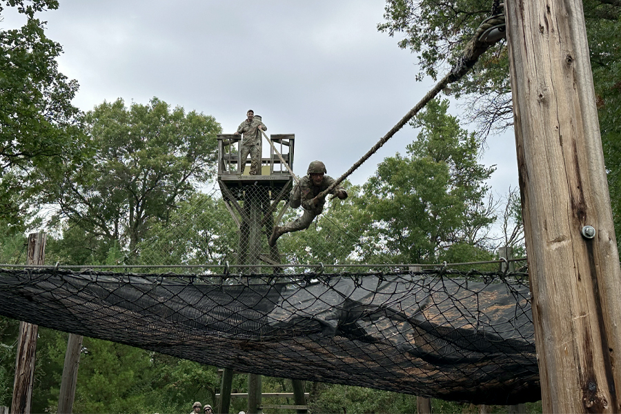 A person dressed in camo fatigues climbs a rope between two wooden posts in the woods while another person looks on.