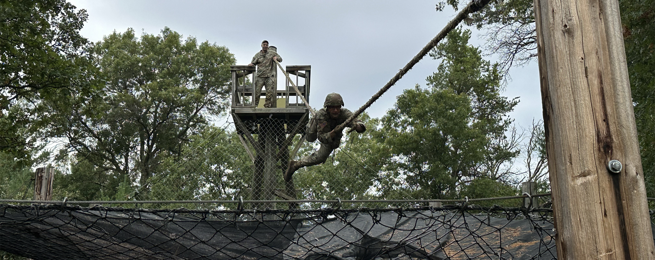 A person dressed in camo fatigues climbs a rope between two wooden posts in the woods while another person looks on.
