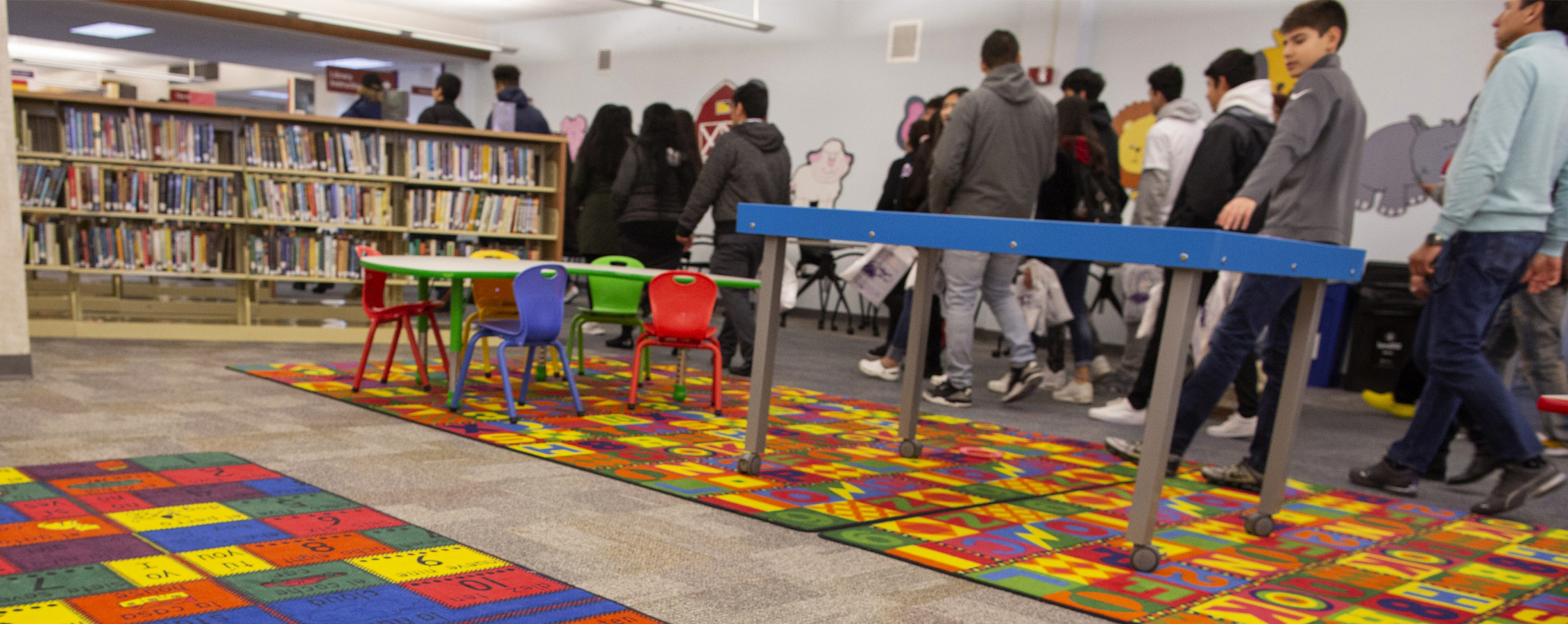 Children walk through a library with bookshelves and colorful rugs.