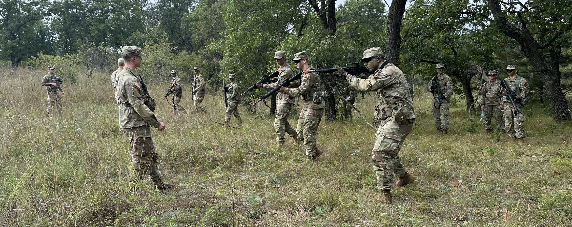 A line of people in camo fatigues walk through a wooded area with rifles in position.