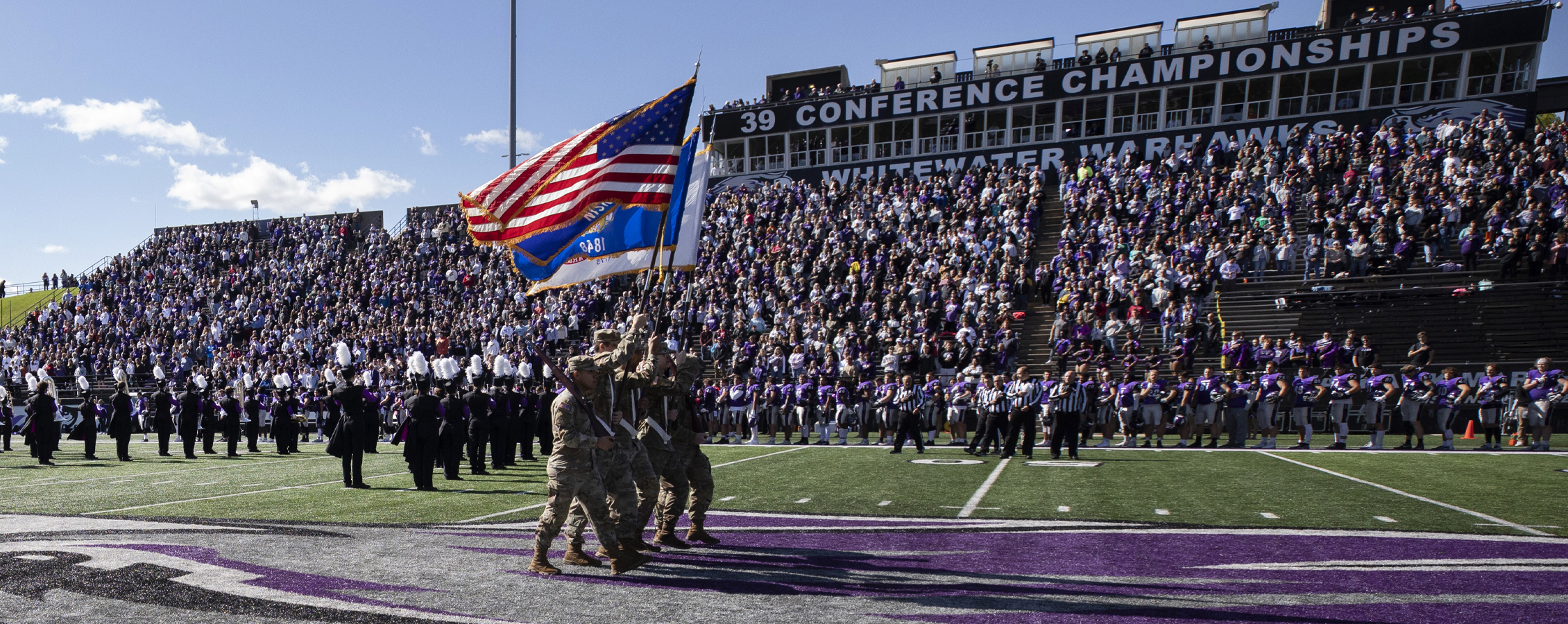 A group of ROTC members dressed in camo fatigues carry the American flag across the football field.