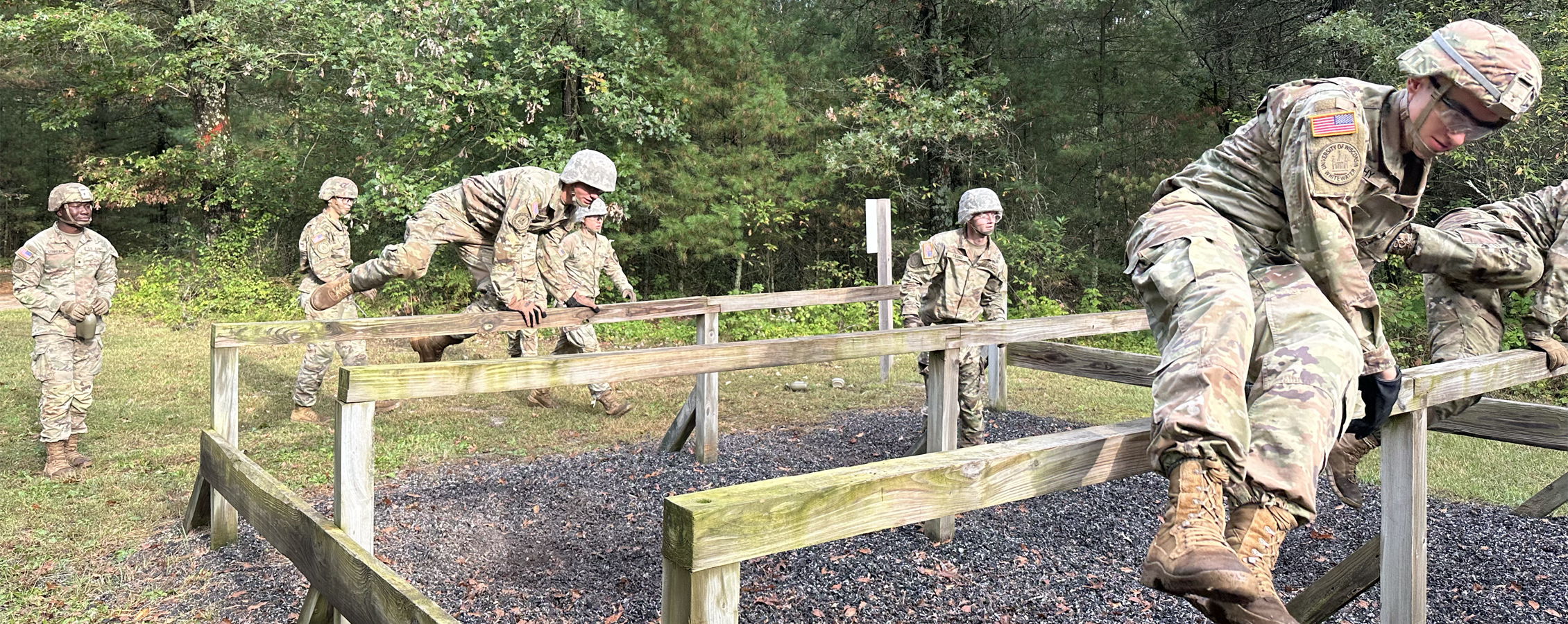 A group of people in camo fatigues and helmets jump over wooden posts as part of an outdoor course.