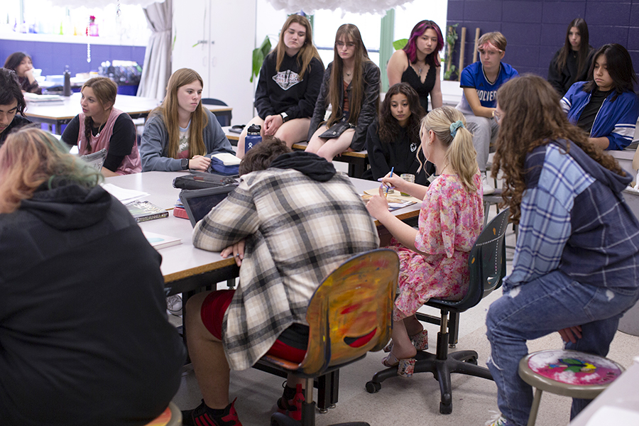 A student teacher sits amongst high schoolers and performs a demonstration on paper.