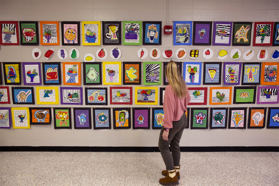 A student teacher stands in front a wall covered with colorful paintings.