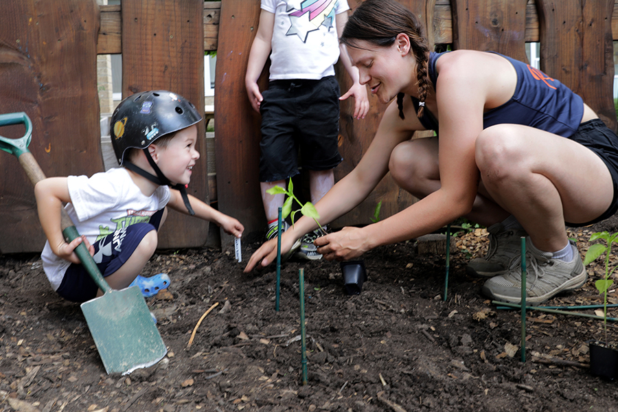 A student and a child kneel on the ground to plant a vegetable in the dirt.