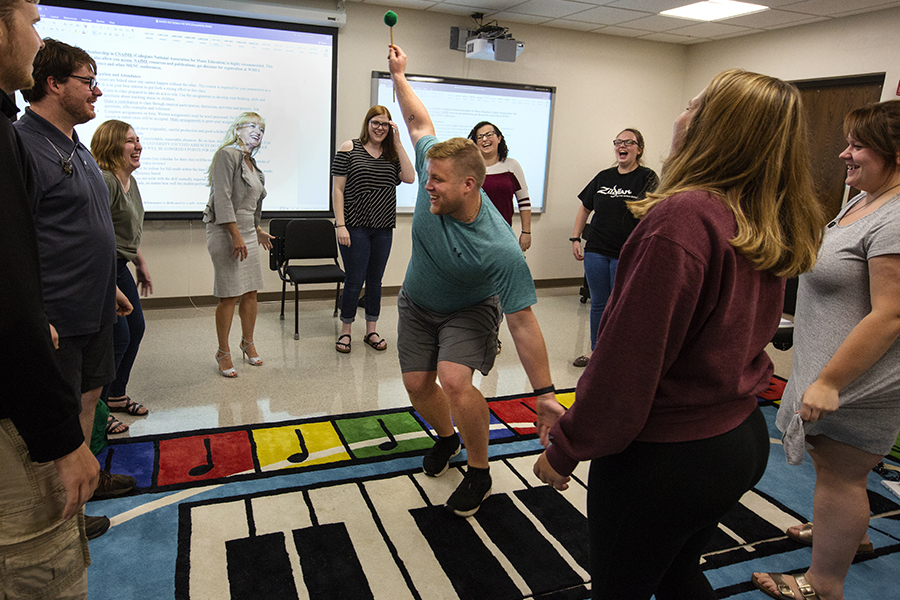 A music teacher talks with their arms raised across a large carpet that looks like a colorful keyboard.