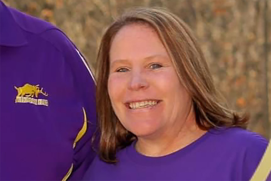 A woman with blonde hair wearing a purple shirt smiles with foliage in the background.