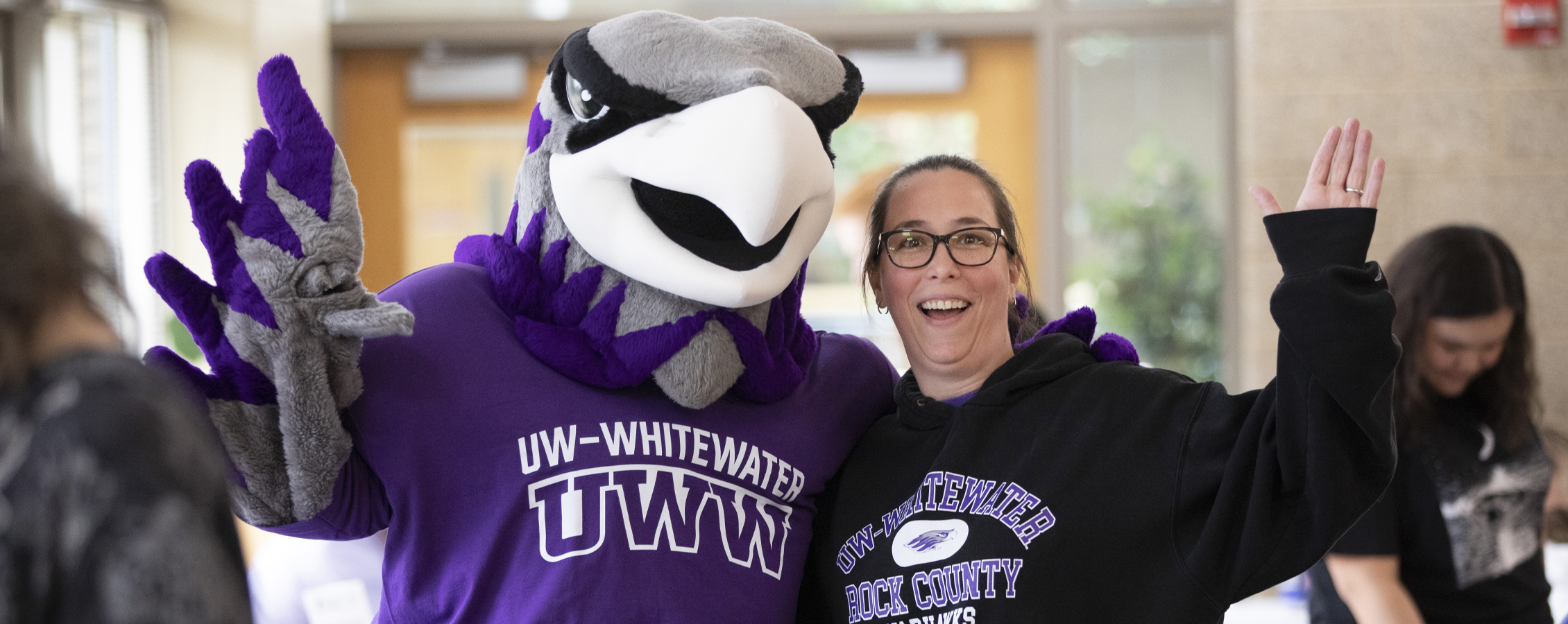 Willie Warhawk stands with a person in a UW-Whitewater sweatshirt and they wave at the camera.