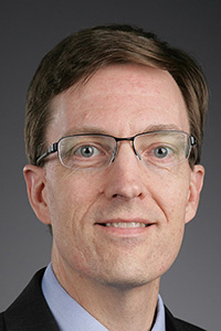 William Ayres wears glasses and a dark suit sitting in front of a dark grey wall.