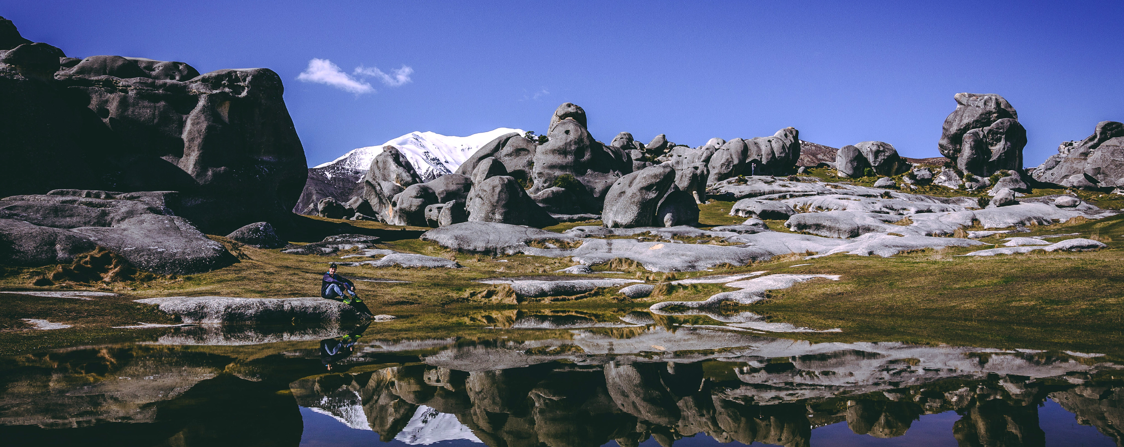 A person sits next to a pool of water amidst trees and mountains.