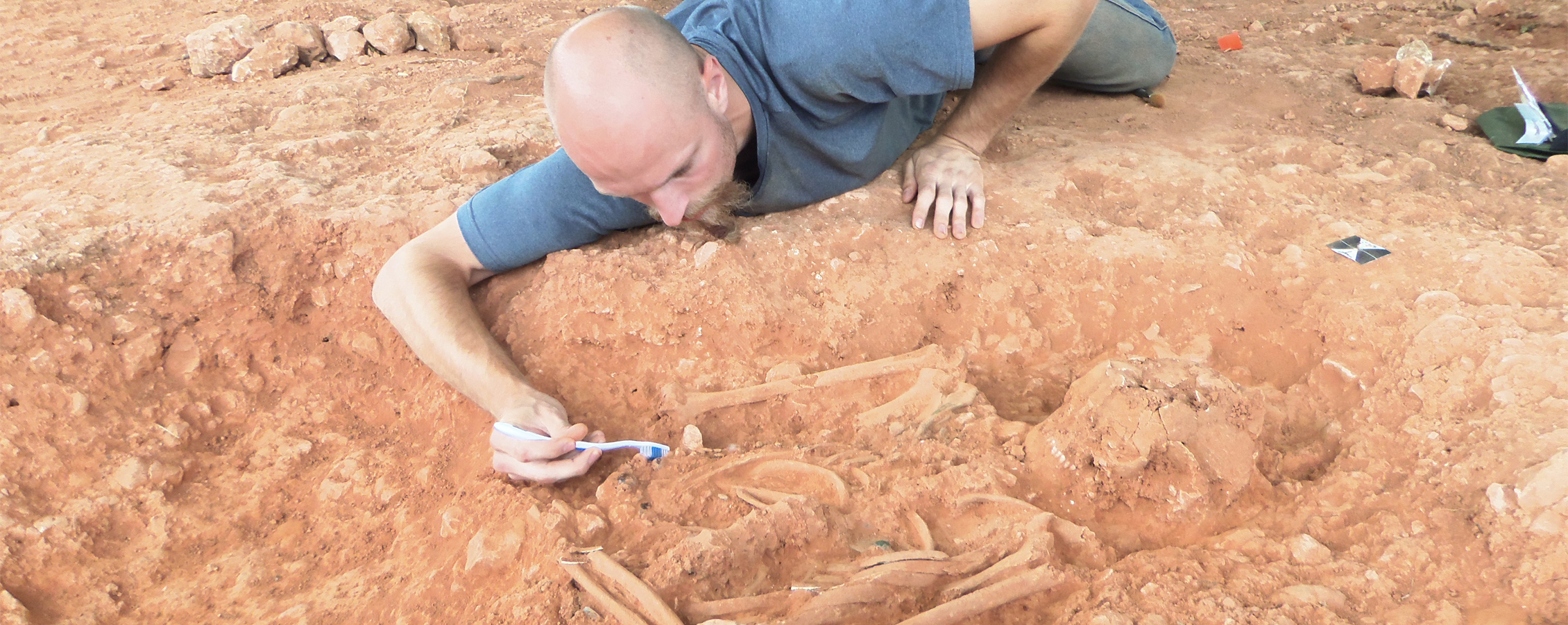 A student lays on the ground and uncovers a skeleton with a toothbrush.