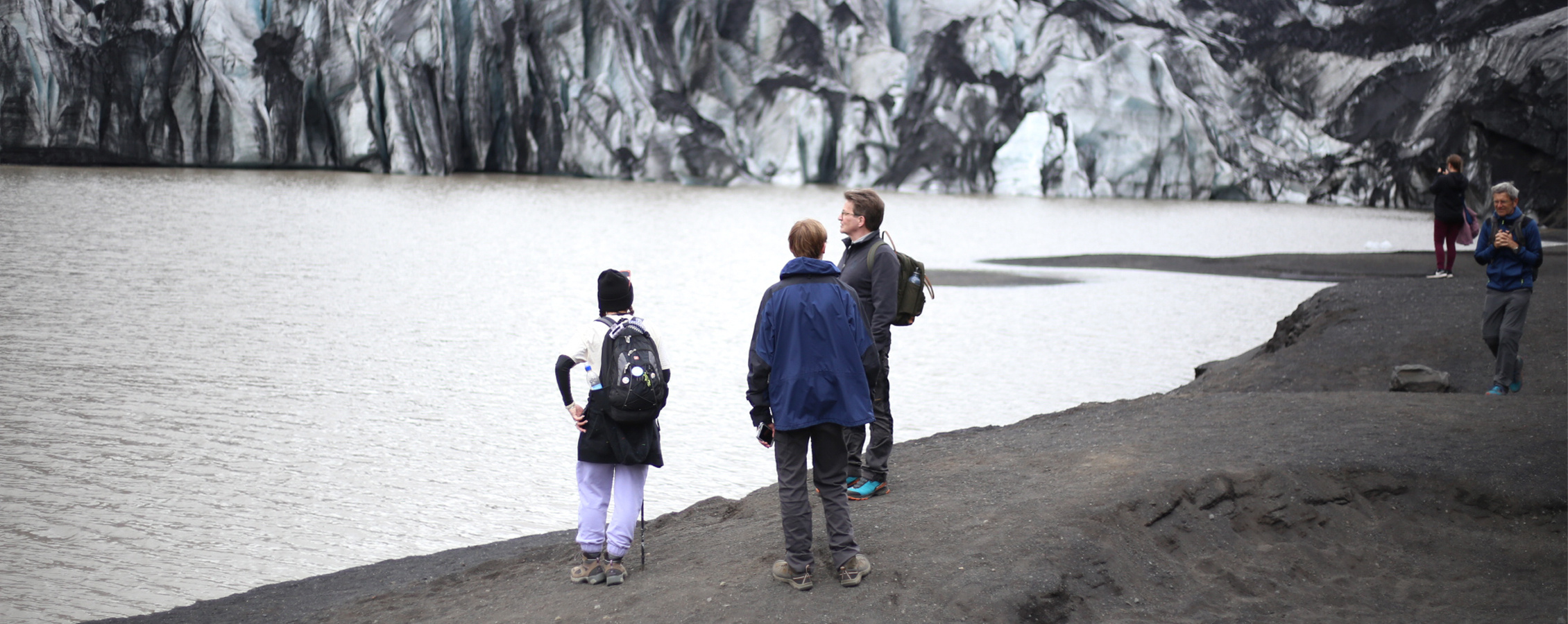 A faculty member stands with students on a shore with snow capped mountains in the background.