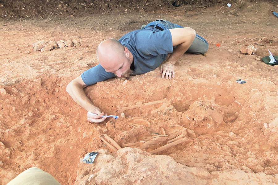 A student lays in the dirt of a dig site and uncovers a skeleton with a toothbrush.