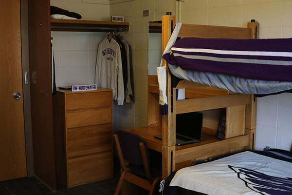 A closet, there is a shelf with a clothes bar, and clothes are hanging off of it. Underneath the shelf there is a dresser.