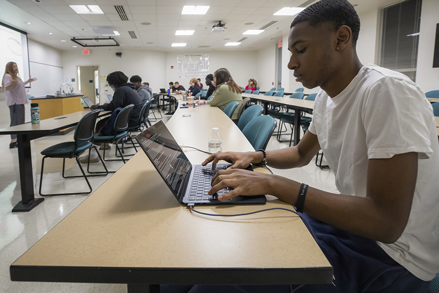 A student sits at a table in a classroom and works on a laptop.