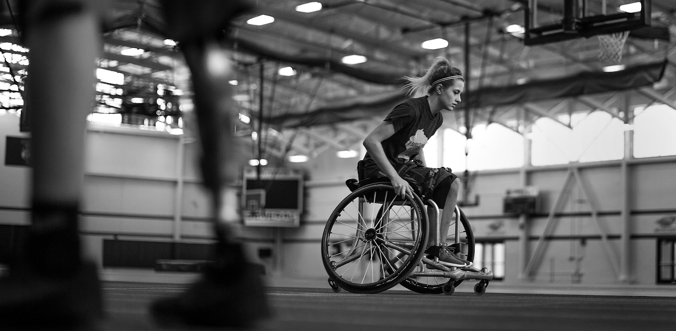 alt="A black and white photo of a person pushing themselves in a wheelchair across the gym floor." 