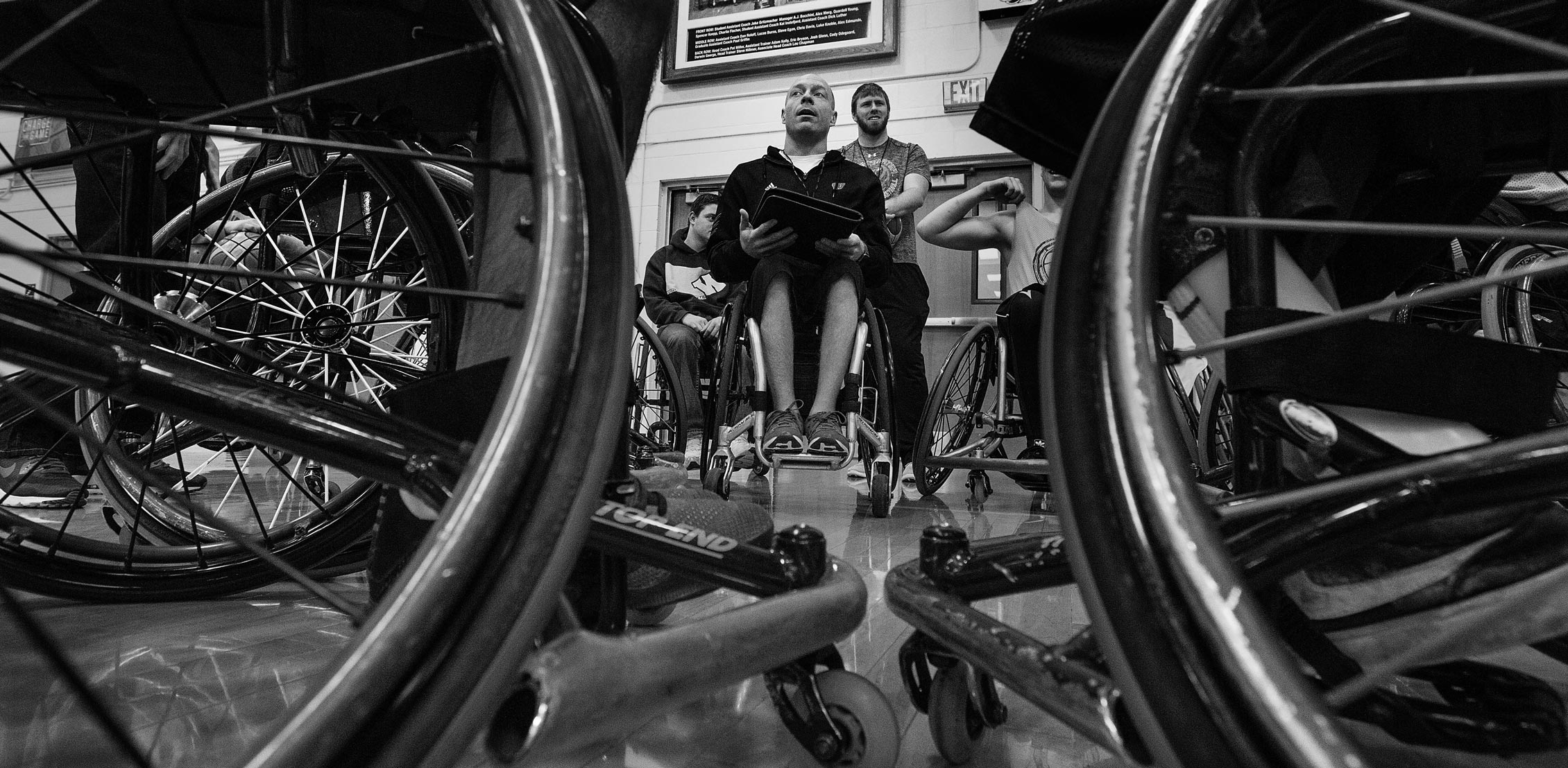 A wheelchair basketball coach is shown in a black and white photo with a clipboard on his lap.