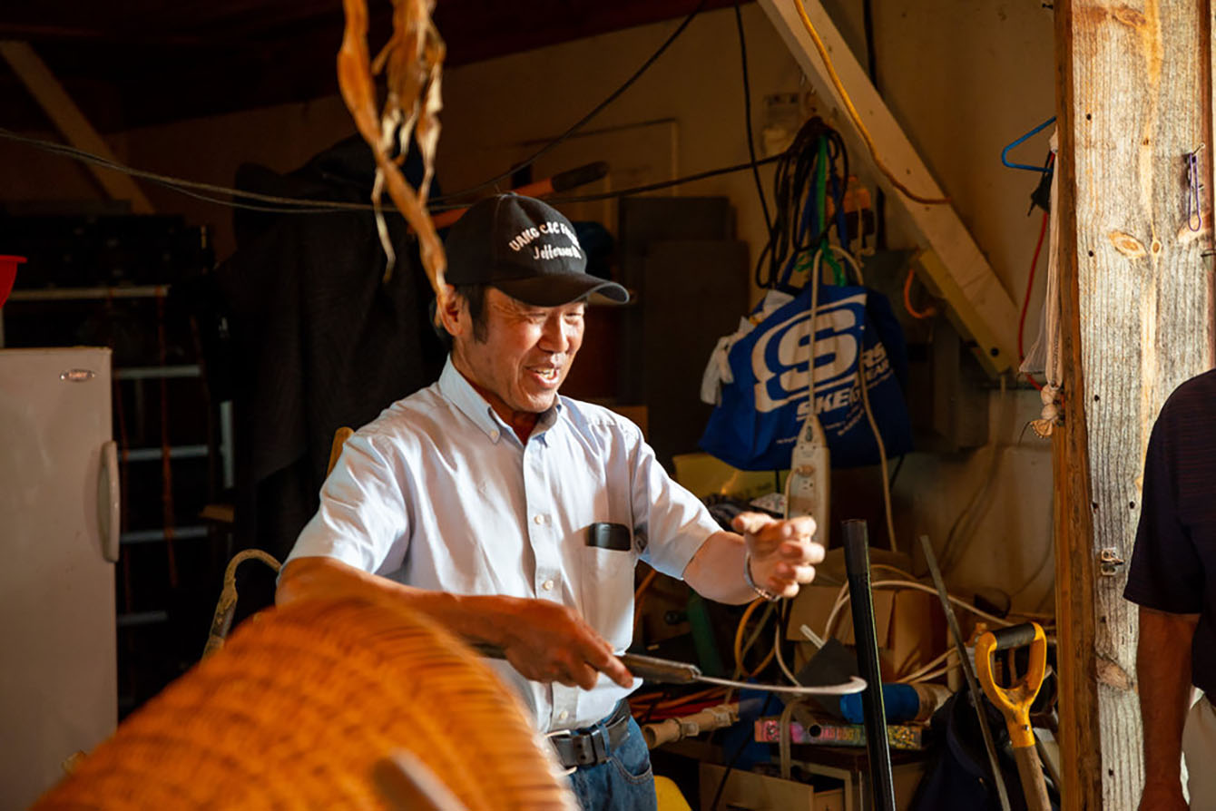 A man stands just outside of a utility shed and demonstrates how to use an old farming tool.