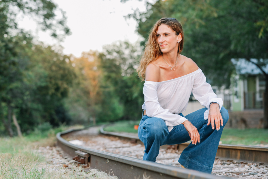 Jen Rulon kneels outdoors on train tracks with a smile on her face and trees in the background.