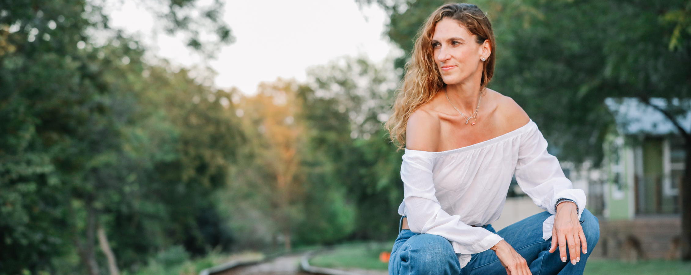 Jen Rulon kneels outdoors with a smile on her face and trees in the background.