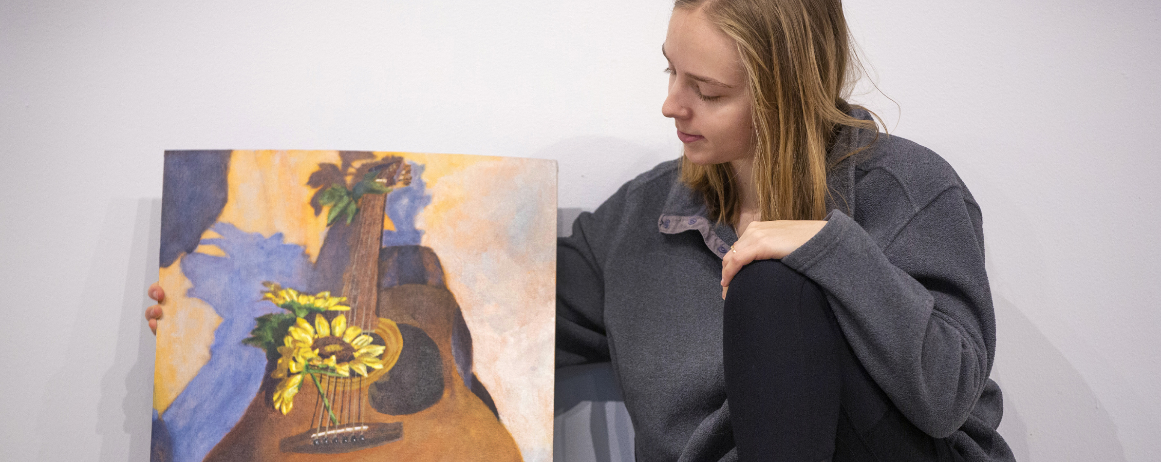 Emma Siskoff sits next to a painting on a guitar.