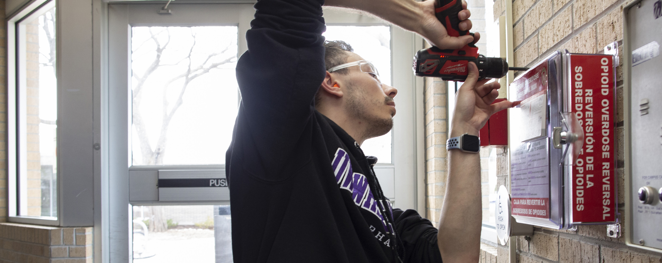 A person uses a drill to install a red opioid overdose reversal box on a brick wall.