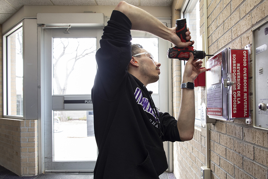 A person uses a drill to install a red opioid overdose reversal box on a brick wall.