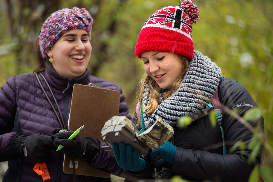 Two women are outside looking at a motion detecting trail camera.