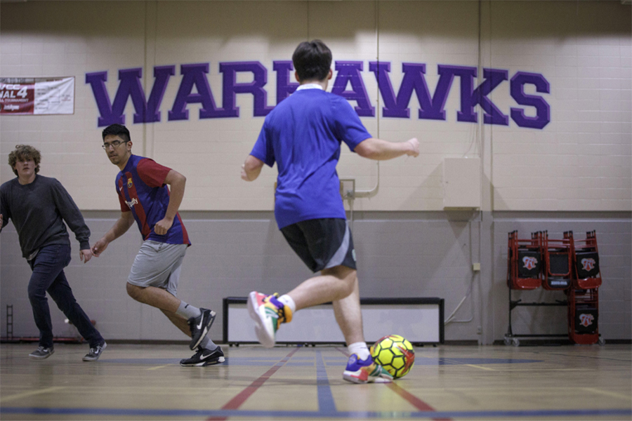 A student dribbles a soccer ball in a gym with the words Warhawks on the wall.