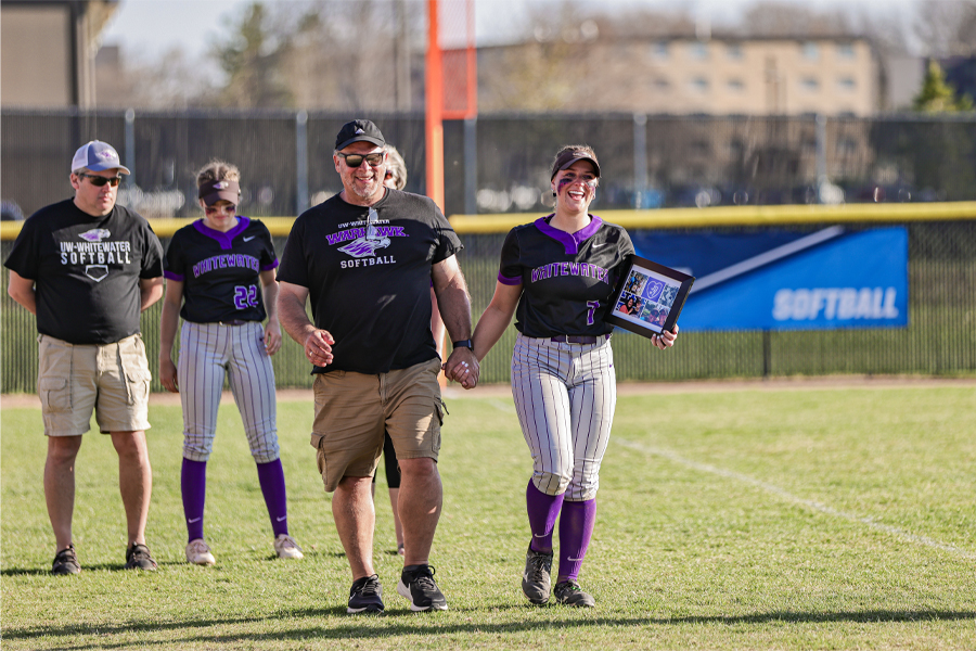 A Warhawk softball player walks hand in hand with her dad on the softball field while smiling and holding a plaque.