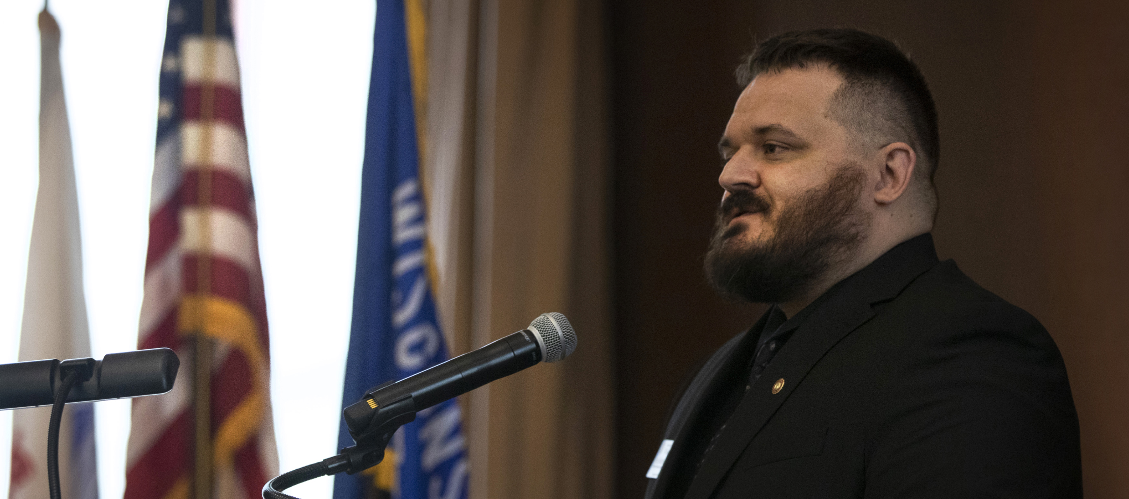 Kris McMenamin stands at a podium with flags hanging in the background.