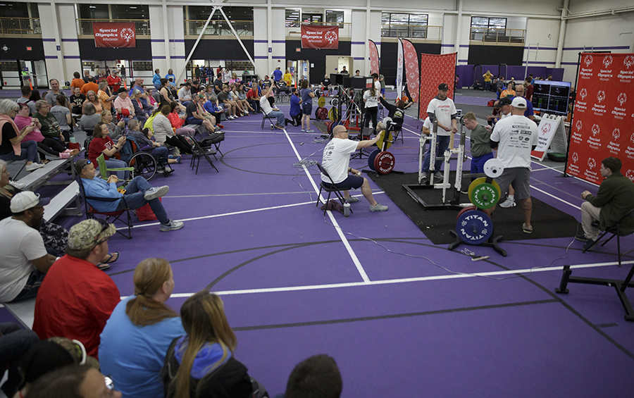 A large group of people gather to watch a Special Olympian weightlifting. 