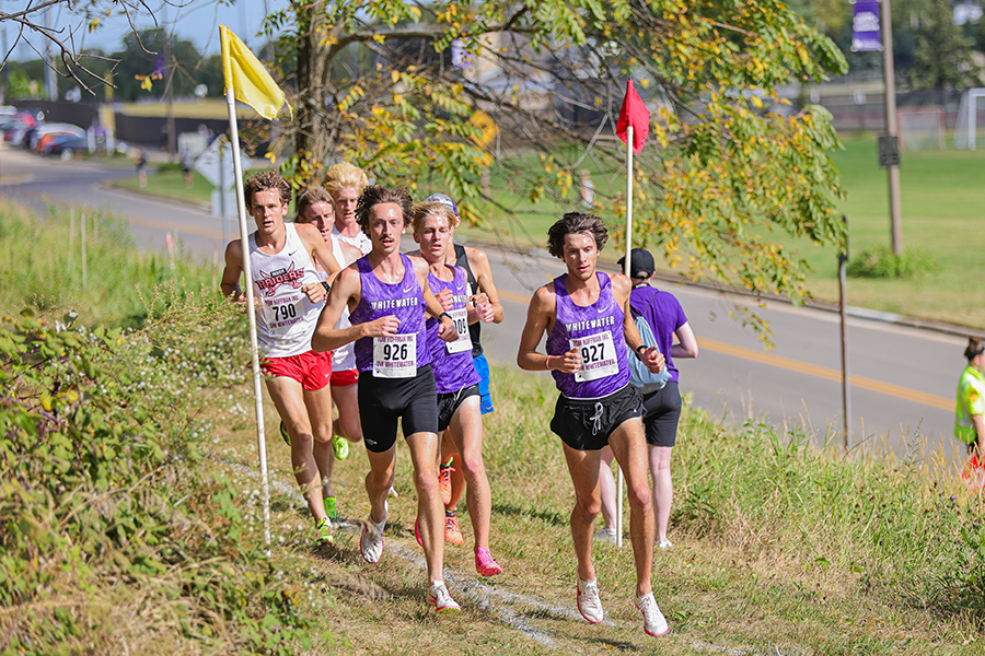 Three Warhawks in purple shirts run in the front of a pack of cross country runners who run by flags on an outdoor trail.