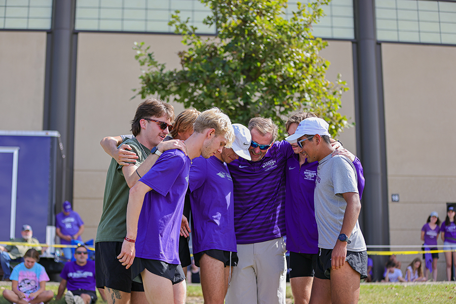 A group of people, wearing mostly purple shirts, group together with smiles on their faces and arms around each other's shoulders. 