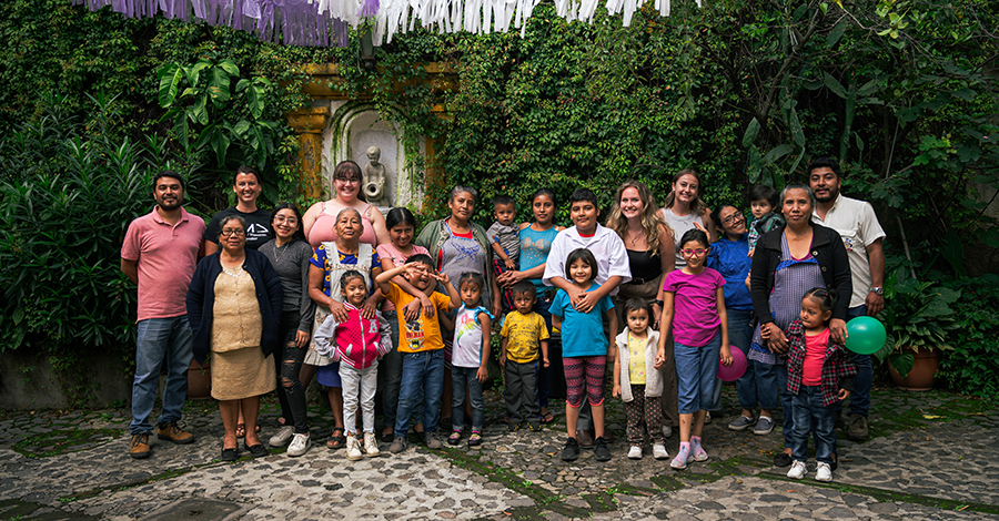 A large group of adults and children stand together in front of a wall partially covered by lush greenery. 