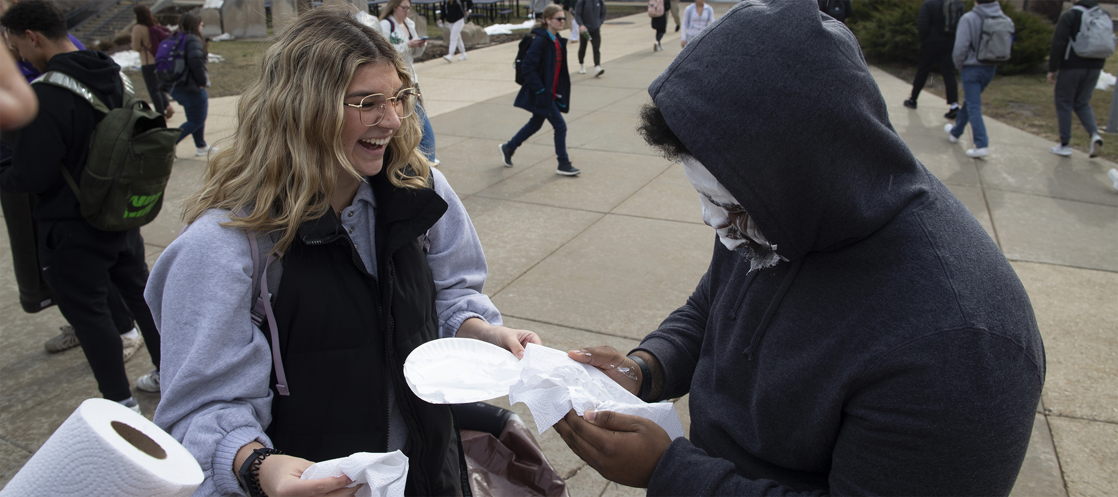 A person smiles and laughs as another person, with pie on their face, holds an empty pie tin.