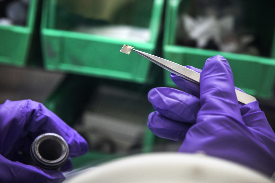 A person wearing purple gloves holds a small piece of metal with tweezers.