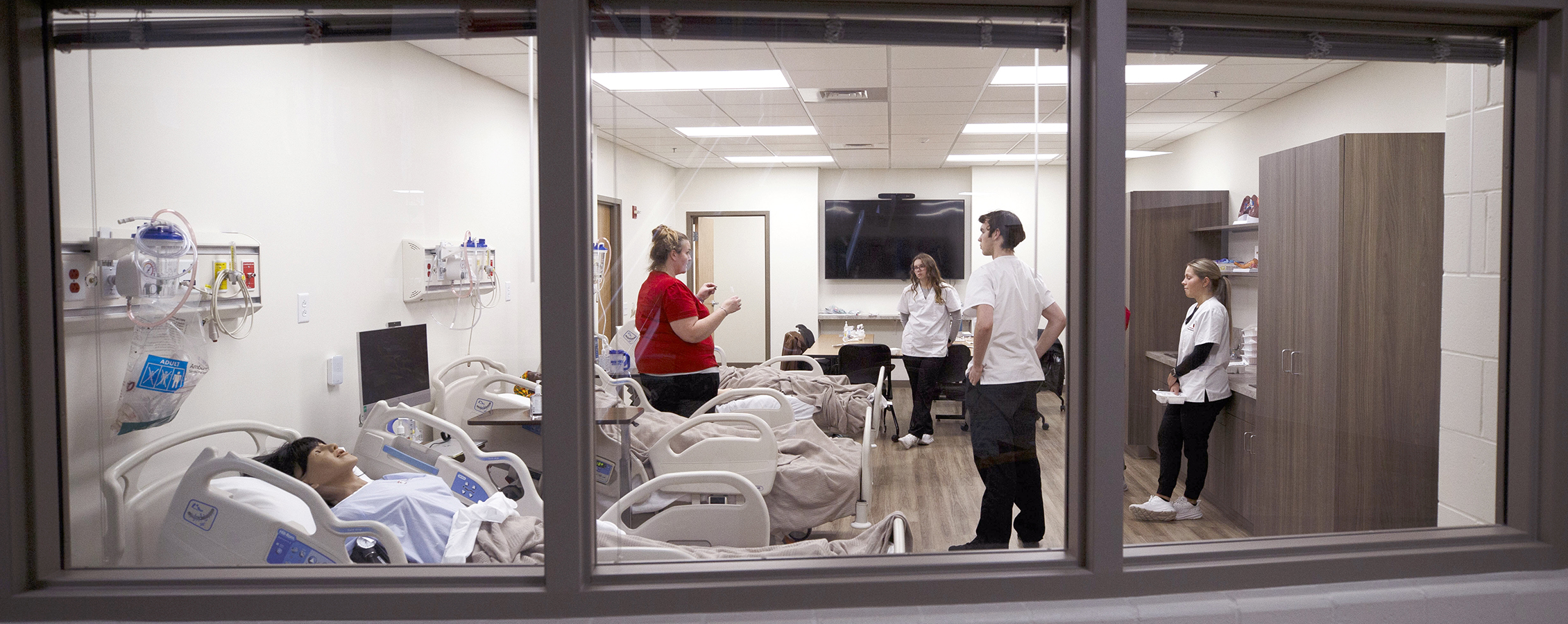 An instructor and three nursing students are seen through a window as they stand in a simulation room with hospital beds.