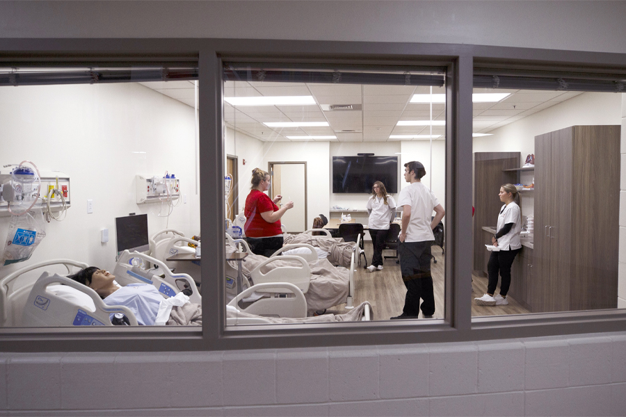 An instructor and three nursing students are seen through a window as they stand in a simulation room with hospital beds.