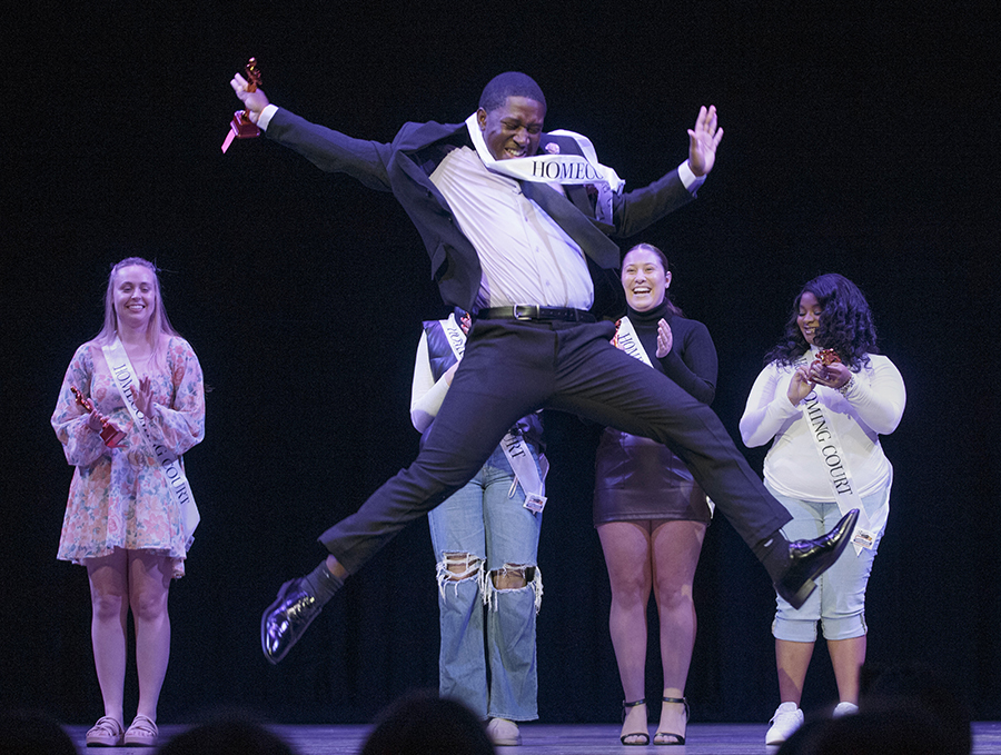 A student on stage wearing a Homecoming banner jumps into the air.