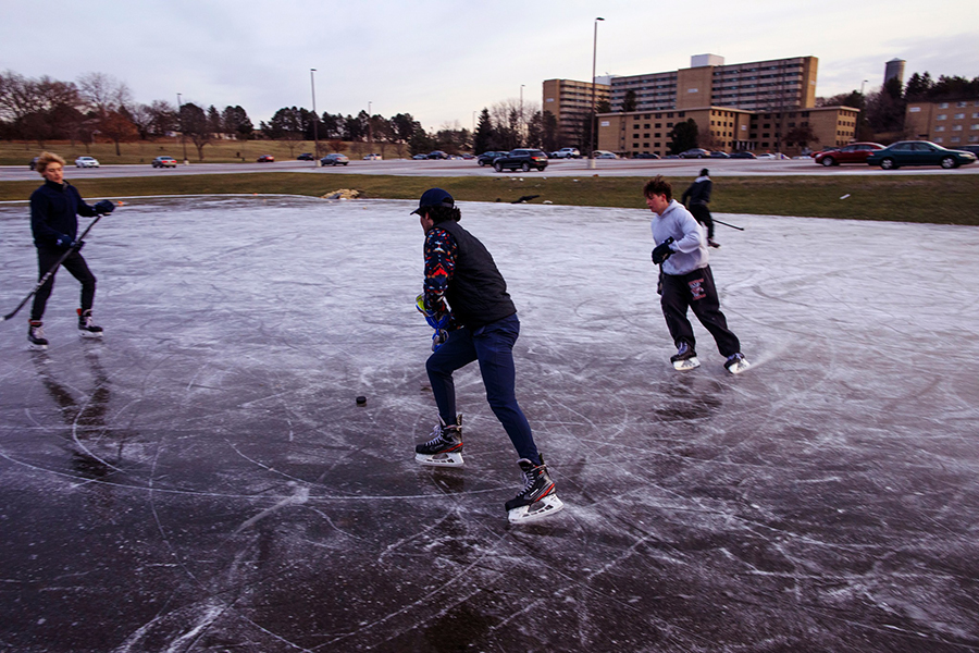 Four students skate on a small pond and play hockey with residence halls in the background.