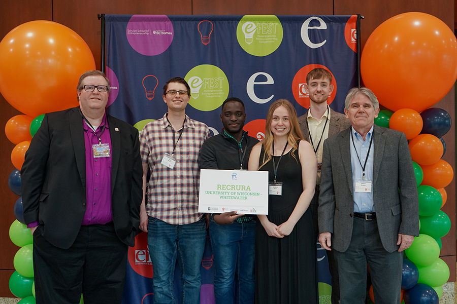 Five people pose for a photo with a plaque and orange balloons behind them.