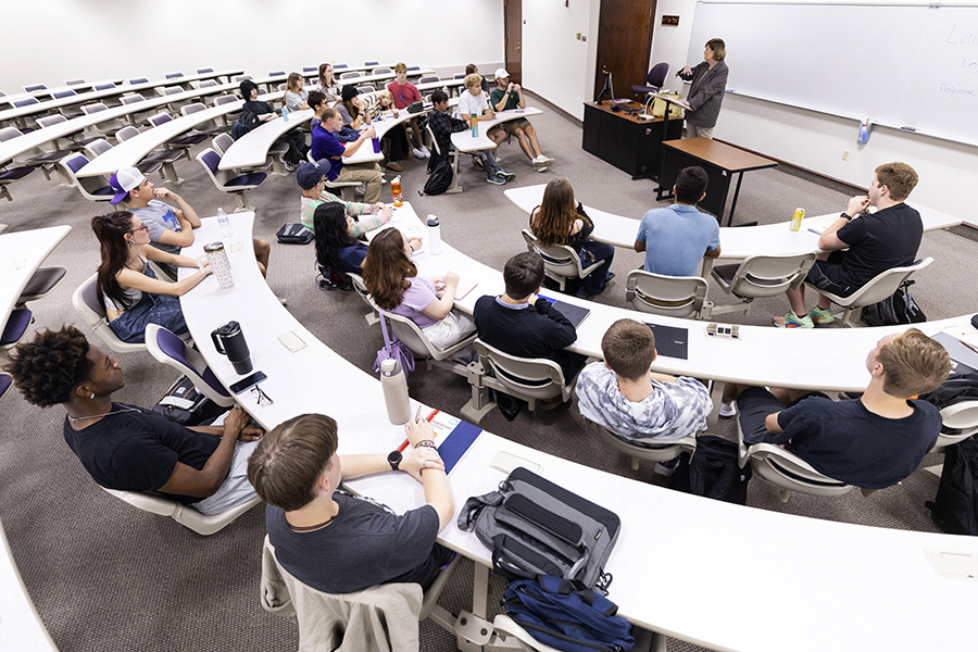 A faculty member teaches at the front of a classroom with students sitting at curved tables.