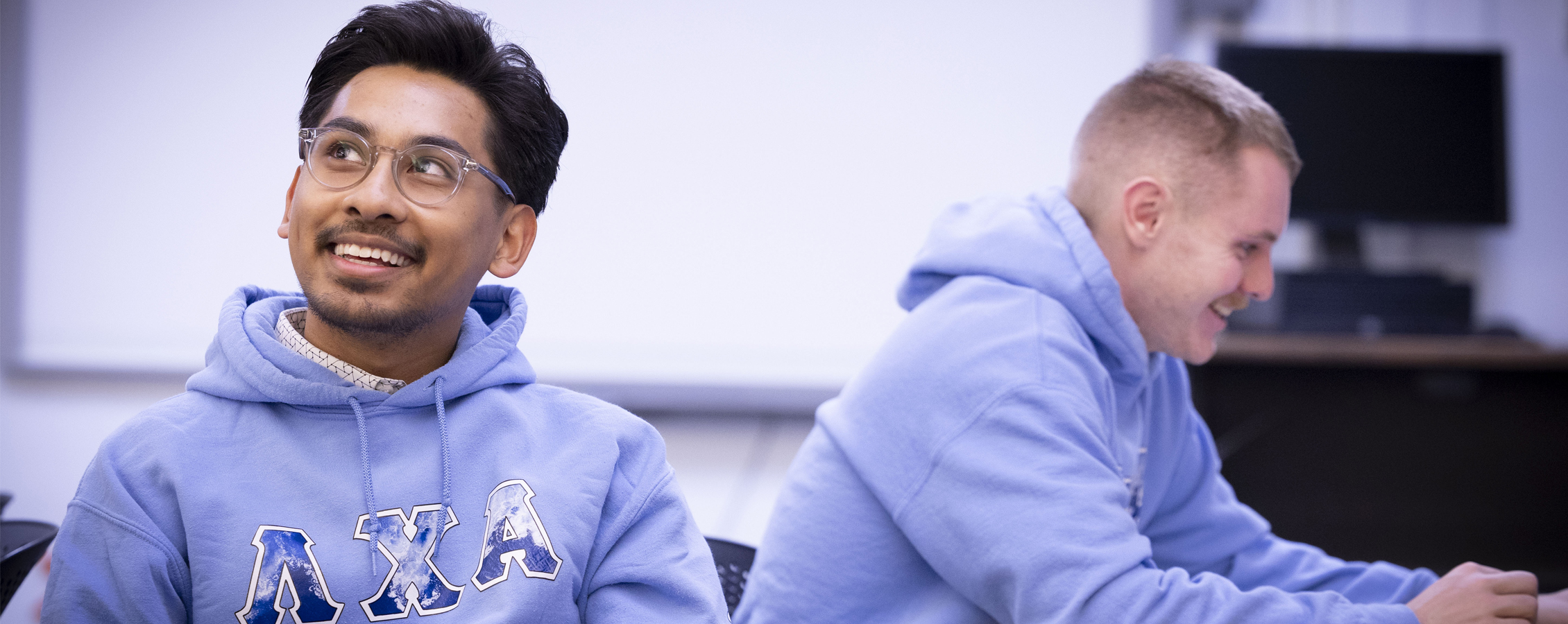 Two smiling men sit at classroom table.