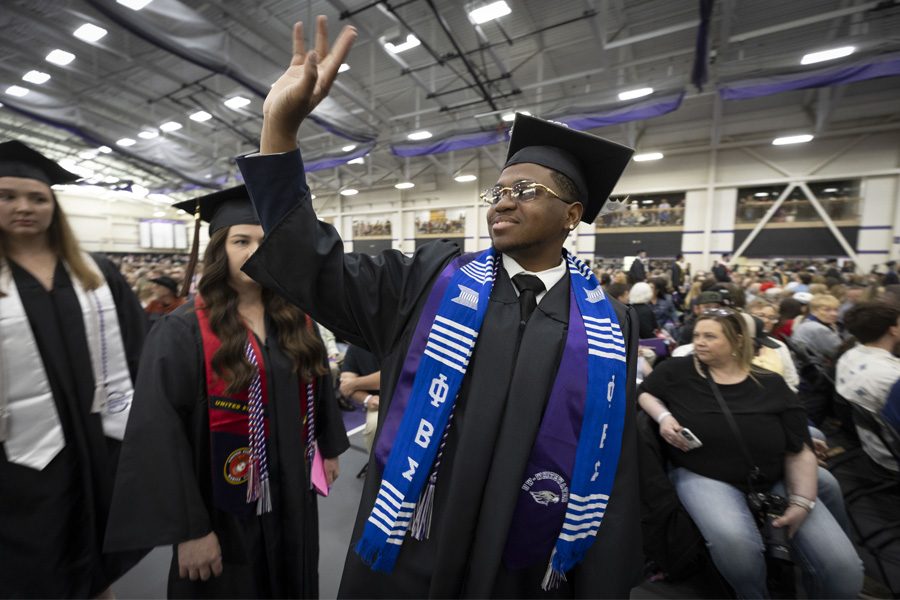 A student in a graduation cap and gown waves to friends and family at commencement ceremony.