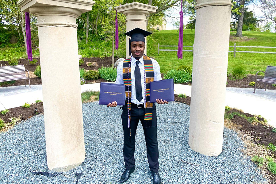 A student wearing a graduation cap holds up two diplomas as they stand in the Memory Garden with three large white pillars.