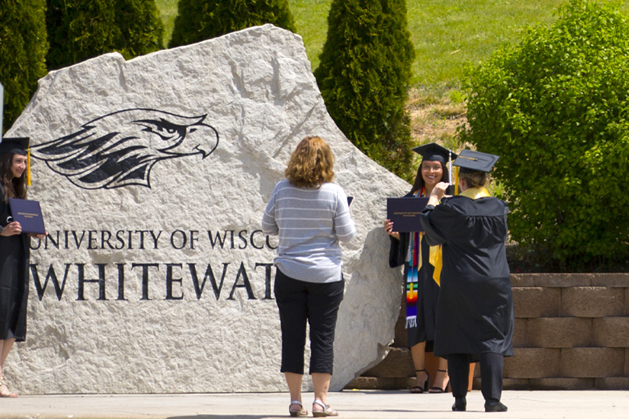 A student wearing graduation regalia stands in front of a large white limestone with UW-Whitewater engraved in it as they get their picture taken.